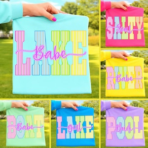 May include: A collection of colorful t-shirts with summer-themed graphics. The shirts feature phrases like "Lake Babe," "Salty Soul," and "Pool Bum" in vibrant colors. The shirts are held up against a green background.