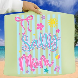 Pu&ograve; includere: Un telo da mare verde chiaro con strisce blu e bianche, con le parole "Salty Mom" in un carattere giocoso. L'asciugamano &egrave; decorato con grafiche a tema spiaggia rosa e gialle, tra cui un fiocco, fiori, stelle, un sole, un pallone da spiaggia e infradito.