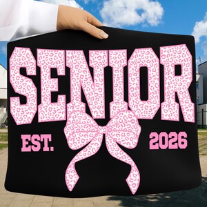 Puede incluir: Cartel negro con la palabra "SENIOR" en letras rosas grandes con estampado de leopardo. Debajo de la palabra "SENIOR" hay un lazo rosa. El texto "EST. 2026" est&aacute; debajo del lazo. El cartel se sostiene contra un cielo azul.