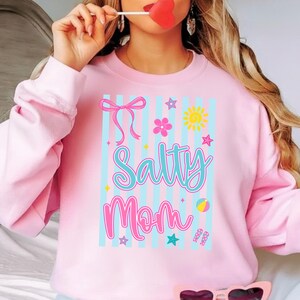 Pu&ograve; includere: Felpa rosa chiaro con la scritta "Salty Mom" in un font fantasioso. Il design include un fiocco, fiori, stelle, un sole, un pallone da spiaggia e infradito. Strisce verticali blu fanno da sfondo.