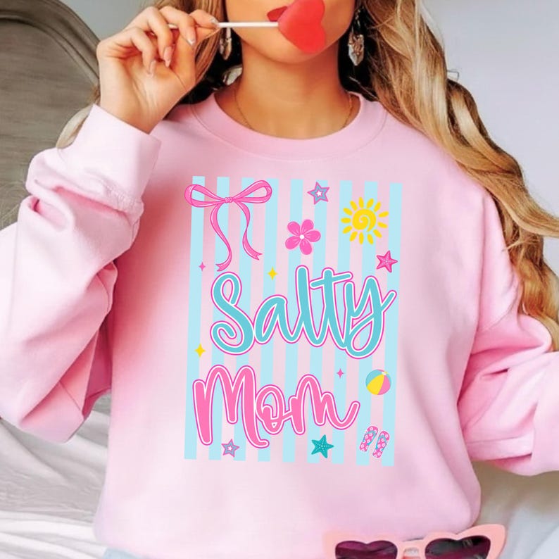 Pu&ograve; includere: Felpa rosa chiaro con la scritta "Salty Mom" in un font fantasioso. Il design include un fiocco, fiori, stelle, un sole, un pallone da spiaggia e infradito. Strisce verticali blu fanno da sfondo.