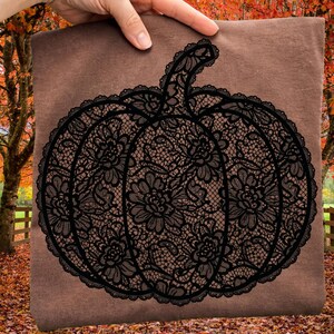 May include: A brown t-shirt featuring a large, black graphic of a pumpkin. The pumpkin design is composed of intricate, detailed black lace patterns with floral motifs, creating a textured, decorative appearance across the center of the shirt.