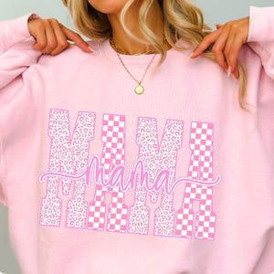 May include: A light pink sweatshirt with the word "MAMA" in a white and pink design. The letters have a mix of leopard print and checkered patterns. The sweatshirt is a casual, comfortable style.