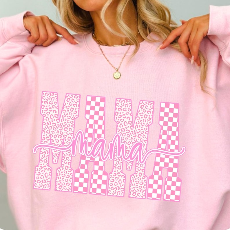 May include: A light pink sweatshirt with the word "MAMA" in a white and pink design. The letters have a mix of leopard print and checkered patterns. The sweatshirt is a casual, comfortable style.
