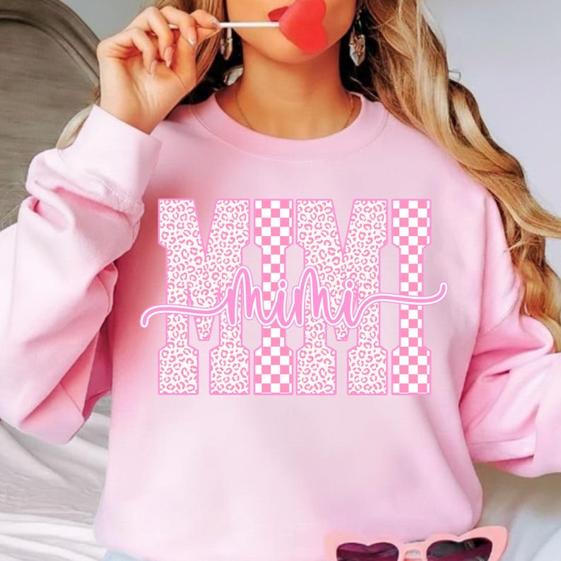 May include: A light pink sweatshirt with the word "Mimi" in a pink script font. The letters "M" are filled with a leopard print pattern, and the "I"s are filled with a pink and white checkered pattern.