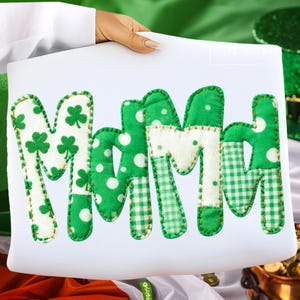 May include: A white rectangular sign displaying the word "MOM" in green patchwork letters. The letters feature shamrocks, polka dots, and gingham patterns. The sign is held by a person.