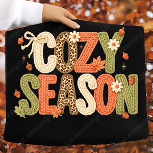 Puede incluir: Cartel rectangular negro con las palabras "COZY SEASON" en un estilo de punto. Las letras son de color crema, naranja, verde y estampado de leopardo. El diseño incluye bellotas, hojas y flores.