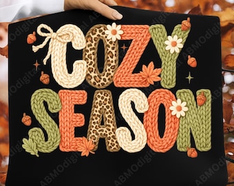 May include: Black rectangular sign with the words "COZY SEASON" in a knitted style. The letters are in cream, orange, green, and leopard print. The design includes acorns, leaves, and flowers.