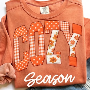 May include: An orange crewneck sweatshirt featuring the word COZY in large block letters with a patchwork design of orange and white polka dots, gingham, and floral patterns. The word Season is printed below in white script. Comfort Colors label.