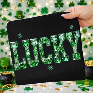 May include: Black clutch with the word "LUCKY" in large, sparkling green gems. The letters are outlined in green and filled with faceted green stones and clear crystals. The clutch is held by a hand.