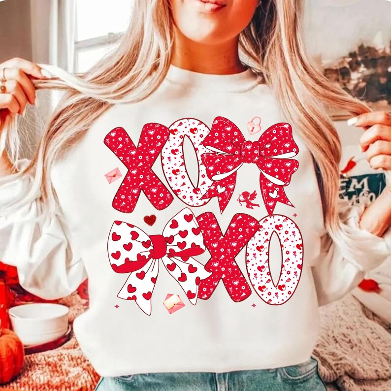 May include: White sweatshirt with a red and white XOXO design. The letters and bows are decorated with hearts and dots. The design includes small hearts, envelopes, and a cupid.