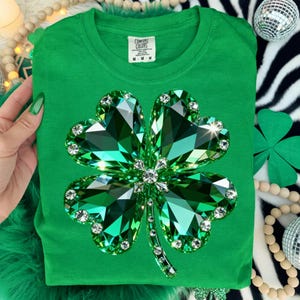 May include: A green t-shirt featuring a large, sparkling four-leaf clover design. The clover is made of faceted green gems with clear crystal accents. The shirt has a crew neck and the Comfort Colors brand tag.