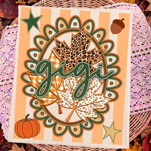 May include: A rectangular sign with a light orange and white striped background featuring the word gigi in dark green script. The design includes a central scalloped oval frame, three maple leaves with leopard, plaid, and leaf patterns, a pumpkin, an acorn, and two stars.