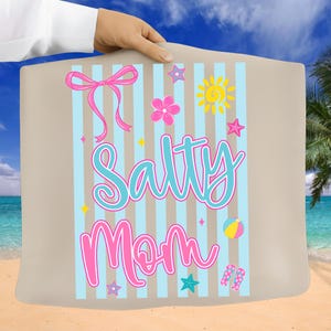 Pu&ograve; includere: Un telo mare beige con strisce verticali blu e la scritta "Salty Mom" in turchese e rosa. L'asciugamano presenta un fiocco rosa, fiori, stelle, un sole, un pallone da spiaggia e infradito.