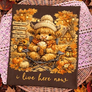 May include: A brown fabric print featuring a fluffy highland cow wearing a knit hat, sitting in a chair and holding a drink. The scene includes pumpkins, lanterns, a house, and signs reading Pumpkin Patch, Apple Orchard, Hayrides, Corn Maze, and i live here now.