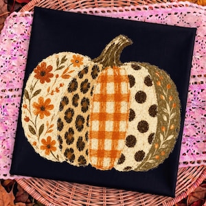 May include: A square black fabric item featuring a central pumpkin illustration. The pumpkin is divided into vertical sections with distinct patterns: orange floral prints, brown leopard spots, an orange plaid, brown polka dots, and leafy vines.