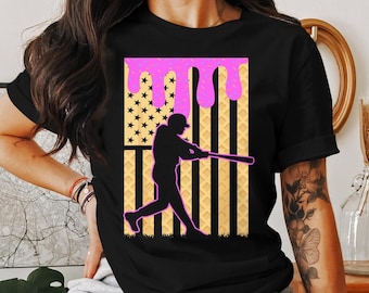 May include: Black t-shirt with a graphic of a baseball player hitting a ball in front of an American flag with pink icing dripping down it.