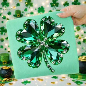 May include: A green square pillow featuring a large, sparkling four-leaf clover design. The clover is made of faceted green gems with clear crystal accents. The pillow is held by a hand, with a St. Patrick's Day theme.