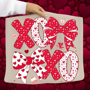 May include: A beige pillow with the letters "XOXO" in red, decorated with heart patterns and bows. The design includes a small cupid, a heart-shaped lock, and tiny envelopes, perfect for Valentine's Day or a romantic gift.