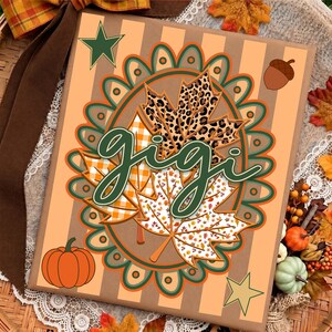 May include: A rectangular sign with a light orange and tan striped background featuring a central scalloped oval frame. Inside the frame are three maple leaves with leopard, orange plaid, and white leaf patterns. The word gigi is written in dark green script.