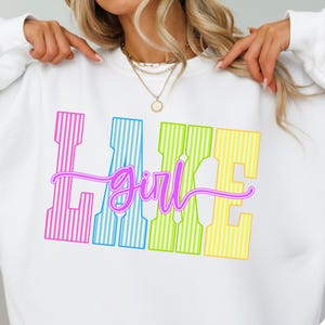 May include: White sweatshirt with the text "Lake Girl" in a colorful, striped design. The word "Lake" is in pink, blue, green, and yellow vertical stripes. The word "Girl" is in a cursive, purple font.