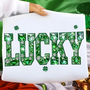 May include: A white sign with the word "LUCKY" spelled out in large, sparkling green gems. The letters are outlined in green and filled with various sizes of green and clear crystals. Small green shamrocks are scattered around the word.