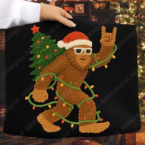 May include: Black tote bag featuring a brown Bigfoot character wearing a Santa hat and sunglasses, carrying a Christmas tree. The Bigfoot is wrapped in Christmas lights and giving a rock-and-roll hand gesture.