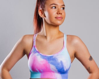 Może przedstawiać: Kobieta w sportowym biustonoszu i dopasowanych legginsach w kolorze różowym, niebieskim i białym z wzorem tie-dye. Sportowy biustonosz ma okrągły dekolt i cienkie ramiączka.