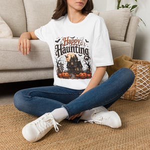 May include: A white short-sleeve t-shirt featuring a graphic print with the text Happy Haunting in a decorative font. The design includes a dark haunted house, a full moon, bare trees, bats, and orange pumpkins on a path with a signpost.