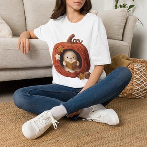 Puede incluir: Una camiseta blanca de manga corta con un gráfico de una calabaza naranja de ganchillo con un pequeño fantasma dentro leyendo un libro. El diseño incluye pequeñas estrellas de ganchillo y una taza, todo con una estética texturizada.