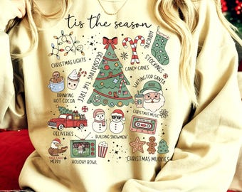 May include: Beige sweatshirt with a festive Christmas-themed graphic. The design includes a decorated tree, candy canes, snowmen, and the phrase "Tis the season." Other elements include Christmas lights, stockings, and a red truck.