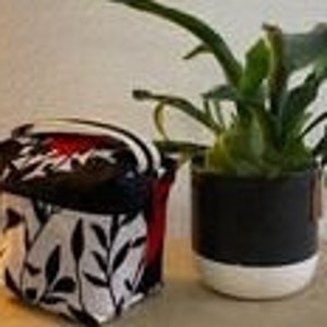 Peut inclure: Une petite pochette zippée à imprimé floral noir et blanc avec un tirette de fermeture éclair rouge et blanche. La pochette est posée sur une surface en bois à côté d'une plante en pot.