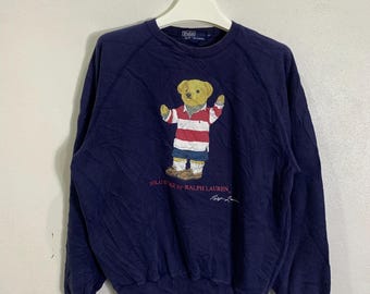 May include: A navy blue long-sleeved sweatshirt with a graphic of a cartoon bear wearing a red and white striped shirt. The text "POLO BEAR by RALPH LAUREN" is printed below the bear. The designer's signature is printed below the text.