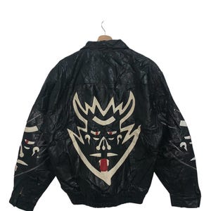 Puede incluir: Chaqueta de cuero negra con un gran diseño de cara de diablo blanco y rojo en la espalda. La chaqueta tiene cuello y cierre de cremallera. Las mangas presentan detalles blancos y rojos adicionales.