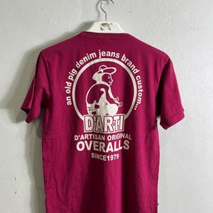 May include: Maroon t-shirt with a white graphic on the back. The graphic features a pig wearing a hat inside a circle with text that reads "an old pig denim jeans brand custom... DARTI D'ARTISAN ORIGINAL OVERALLS SINCE 1979."