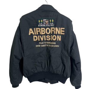 May include: Black bomber jacket with the text "AIRBORNE DIVISION" in tan lettering on the back. The jacket has a patch that reads "FOB TEAM INTERNATIONAL SPECIAL FORCES". The text "PILOT TO BOMBARDER OVER TARGET IN 39% SECONDS!" is also visible.