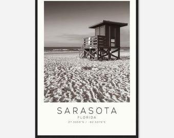 May include: A black and white photograph of a lifeguard stand on a sandy beach. The stand is made of wood and has a roof. The beach is empty and the sand is covered in footprints. The text "SARASOTA FLORIDA 27.3359°N / -82.5379°E" is printed below the image.