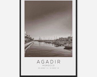 May include: A black and white photograph of a harbor in Agadir, Morocco. The photo shows a row of boats docked in the harbor, with buildings and palm trees in the background. The text "AGADIR MOROCCO 30.4280° N / 9.5925° W" is printed at the bottom of the photo.