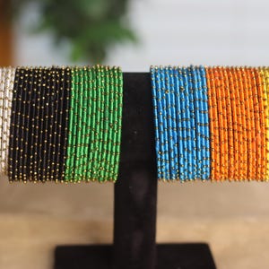 Może przedstawiać: Prezentacja kolorowych bransoletek z koralików. Bransoletki ułożone są w rzędach na czarnym stojaku. Kolory obejmują biały, czarny, zielony, niebieski, pomarańczowy i żółty. Każda bransoletka wykonana jest z małych, cylindrycznych koralików ze złotymi akcentami.