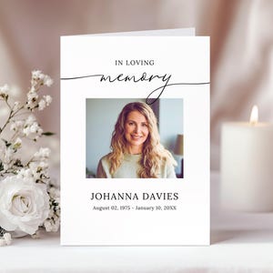 May include: A white memorial card with the words "IN LOVING memory" in black script. Below is a photo of a woman, with the name "JOHANNA DAVIES" and dates of birth and death. A lit candle and white flowers are in the background.