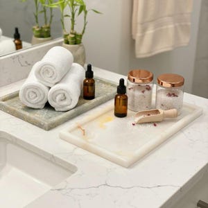Könnte beinhalten: Eine Badezimmer-Arbeitsplattenanordnung mit gerollten weißen Handtüchern, braunen Glasflaschen mit Tropfern und zwei dekorativen Tabletts. Die Tabletts enthalten Gläser mit Kupferdeckeln, Badesalze und einen Holzschöpfer. Grüne Bambuspflanzen sind im Hintergrund.