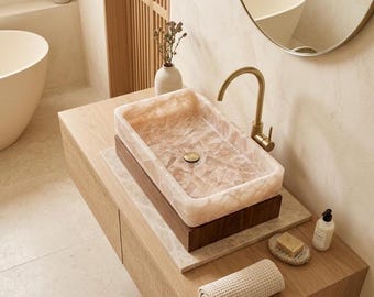 May include: A rectangular, light pink stone sink with a gold tap sits atop a wooden vanity. The sink is mounted on a dark wood base. A rolled white towel, soap, and a bottle are on the vanity.