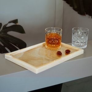Könnte beinhalten: Ein rechteckiges Tablett in hellem Beige mit glatter Oberfläche. Zwei kleine braune Steine und ein Glas mit bernsteinfarbener Flüssigkeit befinden sich auf dem Tablett.