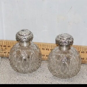 May include: Two clear glass bottles with silver tops and intricate etched designs. The bottles are round and have a decorative pattern.