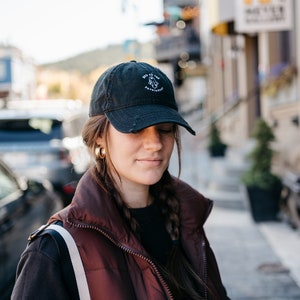 May include: A woman wearing a black baseball cap with the text "Boo to the Patriarchy" embroidered on it. She is wearing a black long-sleeved shirt and a maroon puffer vest.