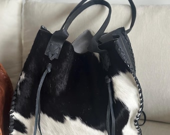 May include: A black and white cowhide tote bag featuring a textured hair-on-hide pattern, two thin black leather shoulder straps, and decorative whipstitch detailing along the side seams. The bag includes a drawstring closure with leather ties.