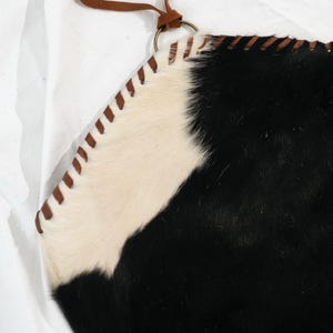 May include: A cowhide bag with a black and white pattern. The bag has a brown leather strap and a metal ring. The edges are stitched with brown thread. The bag is made of genuine cowhide.