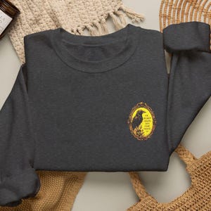 May include: A dark charcoal grey crewneck sweatshirt featuring a small, oval-shaped graphic on the left chest. The graphic depicts a black crow inside an ornate gold frame with the text I'M SILENTLY JUDGING YOUR PLANT CHOICES.
