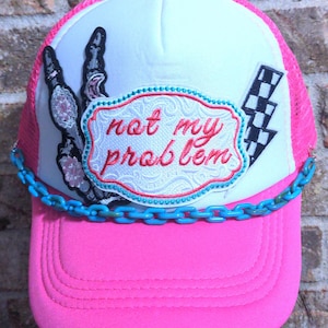 May include: A pink trucker hat with a white front panel and pink mesh back. The hat features an embroidered patch with the phrase "not my problem" and a peace sign. A blue chain is attached to the brim. A black and white checkered flag is also on the hat.