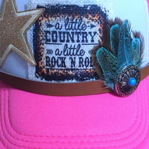 May include: A pink trucker hat with a white front panel and brown brim. The hat features a gold glitter star, a patch with the text "a little COUNTRY a little ROCK N ROLL", and blue feathers with a turquoise center.